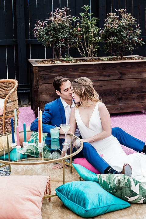 bride and groom sitting on outside furniture pillows, bride is in a formfitting gown with straps and her hair down in a loose style and holding a bright colored bouquet of flowers, while the groom is in a cobalt blue suit with brown shoes and his shirt undone