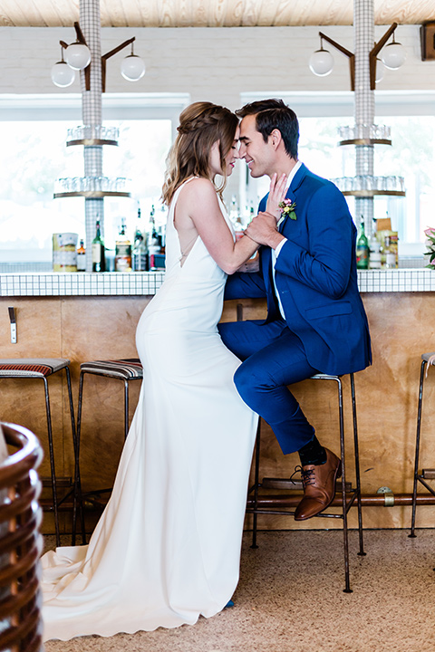 bride and groom sitting at bar, bride is in a formfitting gown with straps and her hair down in a loose style, while the groom is in a cobalt blue suit with brown shoes and his shirt undone