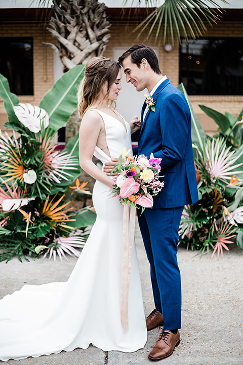 bride and groom standing with their heads touching, bride is in a formfitting gown with straps and her hair down in a loose style, while the groom is in a cobalt blue suit with brown shoes and his shirt undone