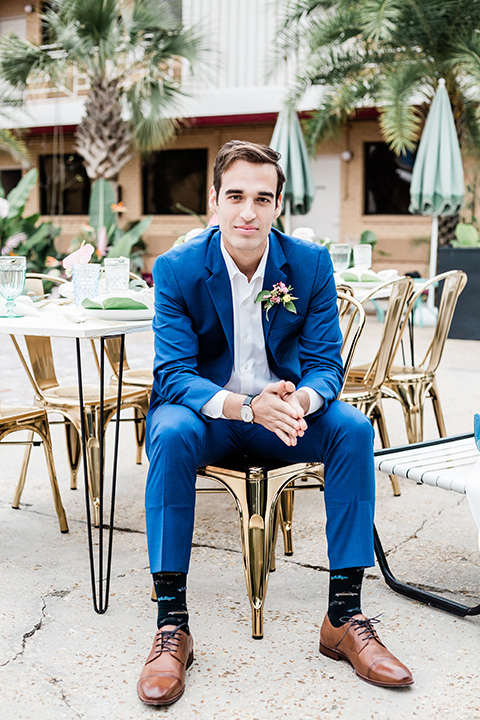 the groom is in a cobalt blue suit with brown shoes and his shirt undone sitting next to the pool