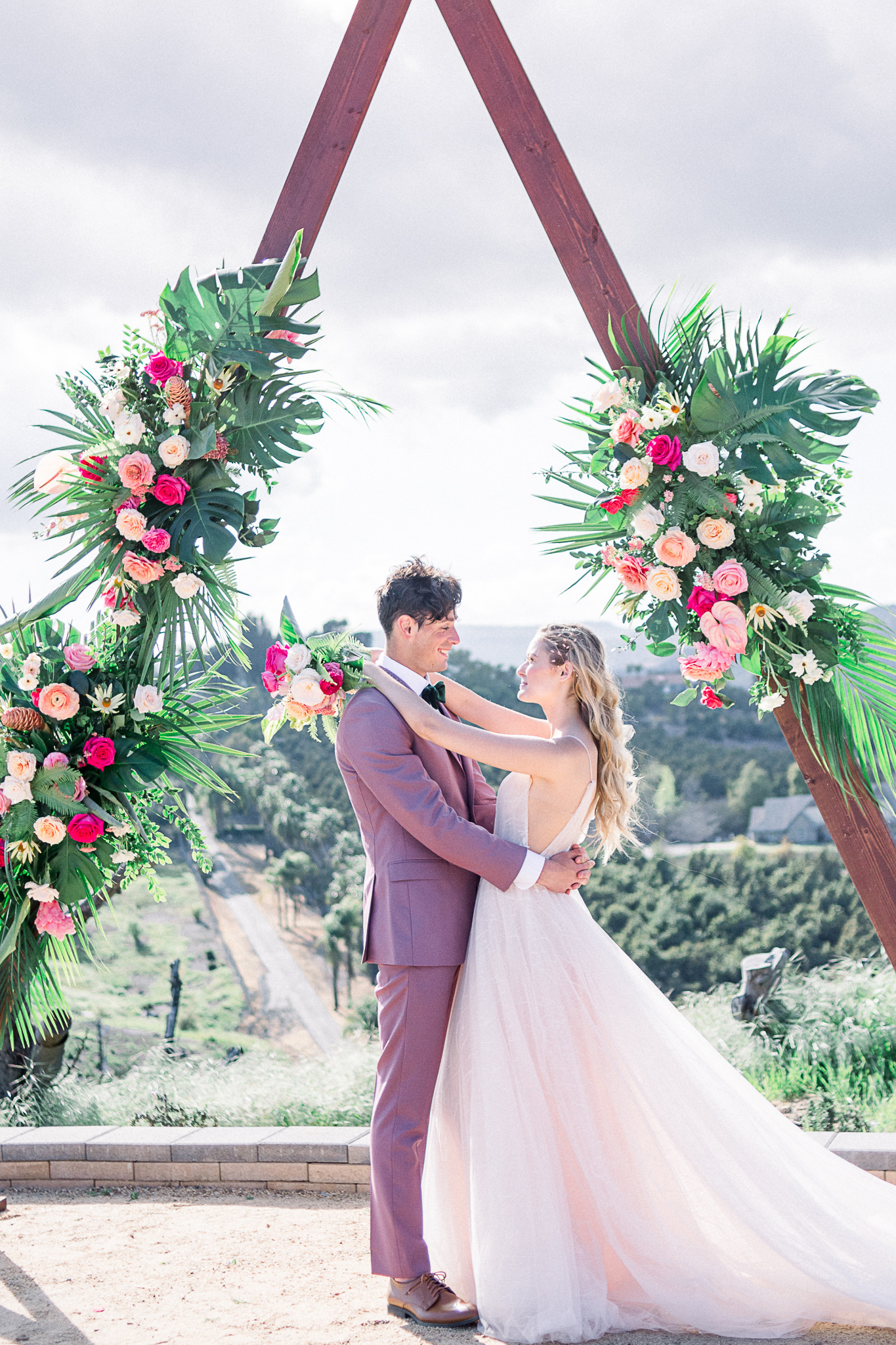 emerald-peak-temecula-wedding-bride-and-groom-looking-at-each-other-at-ceremony-arch-bride-in-a-blush-toned-tulle-ballgown-with-straps-groom-in-a-rose-pink-suit-with-a-green-velvet-bow-tie