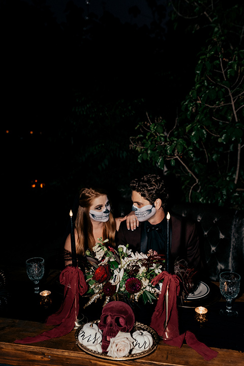  bride in a black gown with a black train and groom in a burgundy tuxedo with a black shirt both with their face painted in a skull pattern 