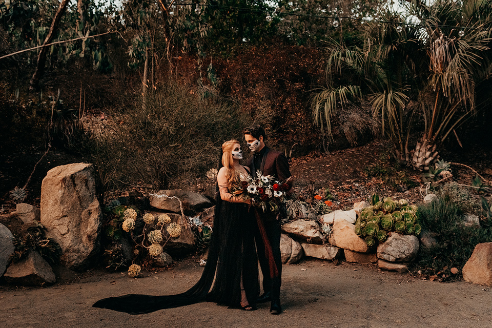  bride in a black gown with a black train and groom in a burgundy tuxedo with a black shirt both with their face painted in a skull pattern 