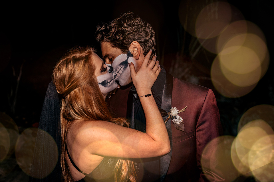  bride in a black gown with a black train and groom in a burgundy tuxedo with a black shirt both with their face painted in a skull pattern 