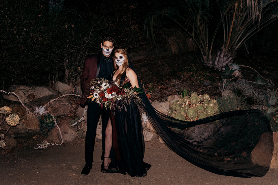  bride in a black gown with a black train and groom in a burgundy tuxedo with a black shirt both with their face painted in a skull pattern 