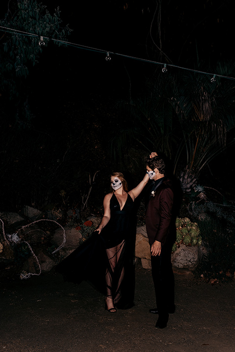 bride in a black gown with a black train and groom in a burgundy tuxedo with a black shirt both with their face painted in a skull pattern