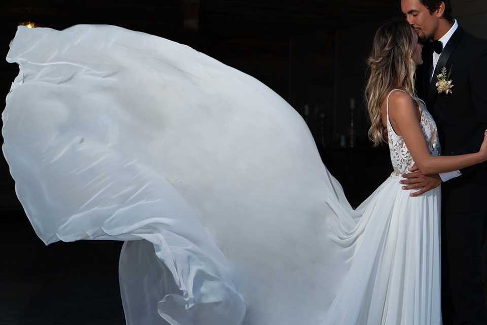 la-bonita-ranch-rustic-shoot-bride-and-groom-brides-dress-blowing-the-bride-in-a-white-bohemian-gown-with-a-nude-underlay-and-lace-detailing-groom-in-a-navy-shawl-lapel-tuxedo-with-a-black-bow-tie