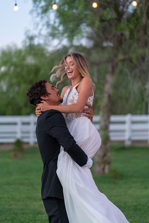 la-bonita-ranch-rustic-shoot-groom-holding-the-bride-up-smiling-bride-in-a-white-bohemian-gown-with-a-high-neckline-with-a-lace-groom-in-a-navy-shawl-lapel-tuxedo-with-a-black-bowtie