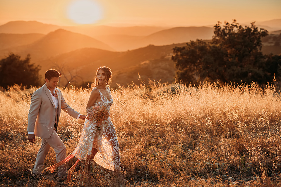  bride in a white bohemian style lace gown with a straps and a her hair in loose waves with the groom in a light grey suit