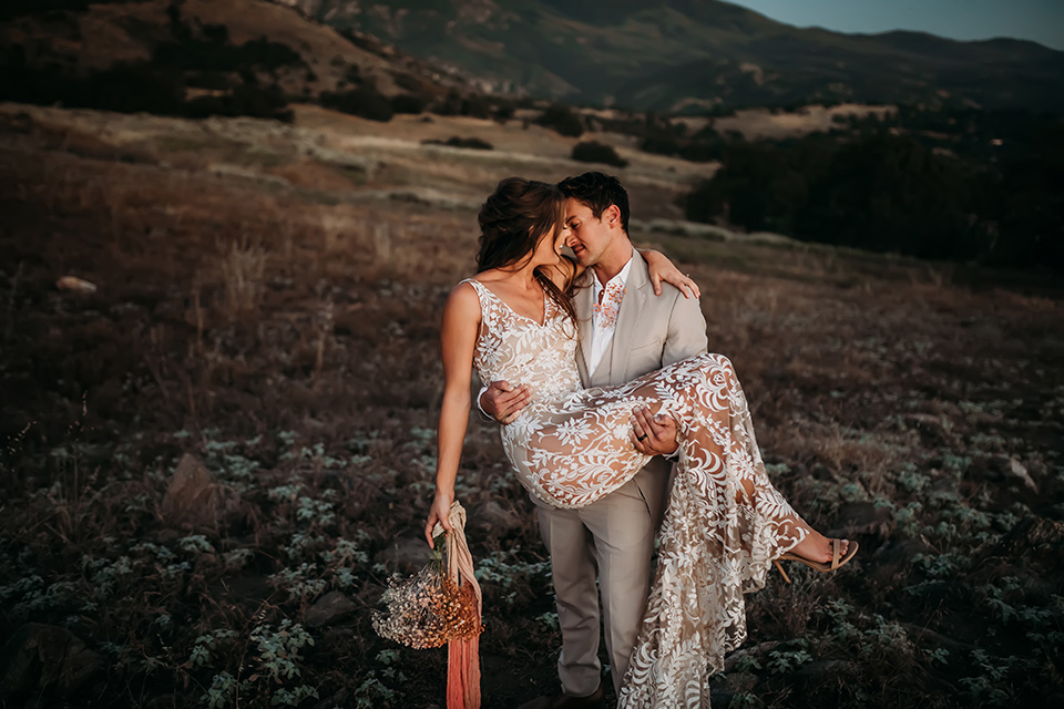  bride in a white bohemian style lace gown with a straps and a her hair in loose waves with the groom in a light grey suit