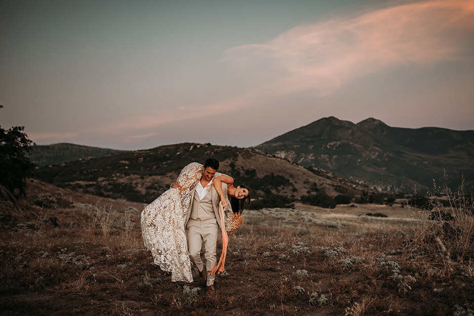  bride in a white bohemian style lace gown with a straps and a her hair in loose waves with the groom in a light grey suit 