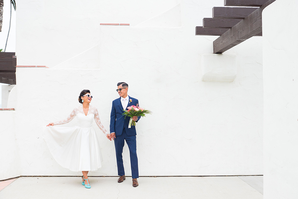 Ole-Hanson-Beach-Club-bride-and-groom-by-white-wall-the-bride-wore-a-tea-length-gown-with-lace-sleeves-and-an-elbow-length-veil-the-groom-wore-a-dark-navy-blue-suit-with-a-blush-pink-long-tie-and-brown-shoes