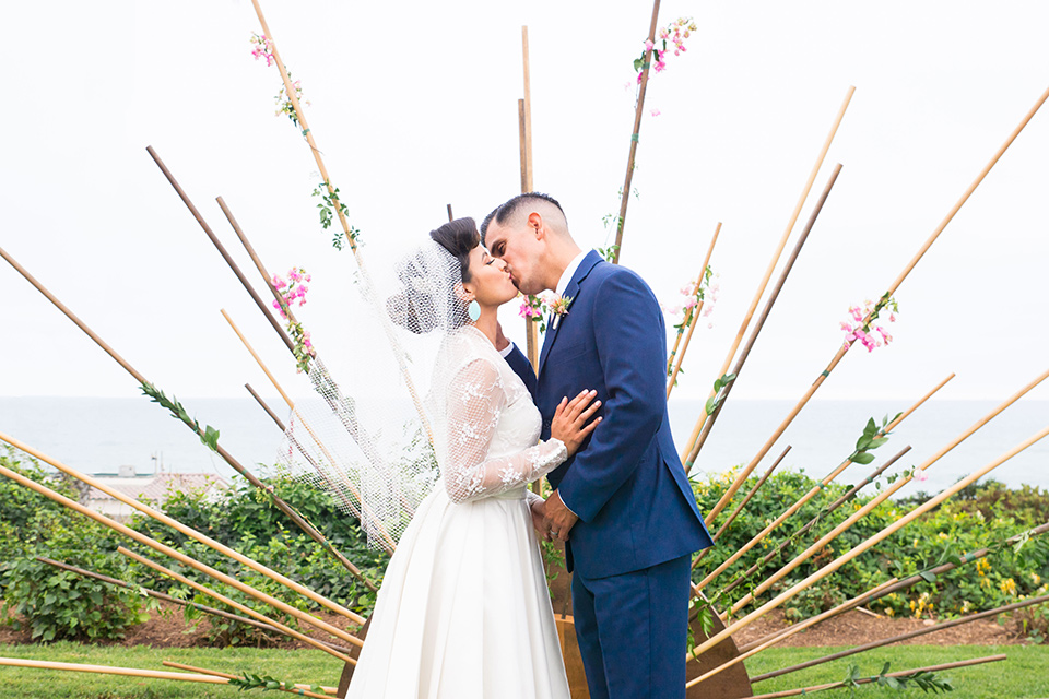 Ole-Hanson-Beach-Club-bride-and-groom-kissing-at-ceremony-the-bride-wore-a-tea-length-gown-with-lace-sleeves-and-an-elbow-length-veil-and-team-retro-heels-the-groom-wore-a-dark-navy-blue-suit-with-a-blush-pink-long-tie-and-brown-shoes