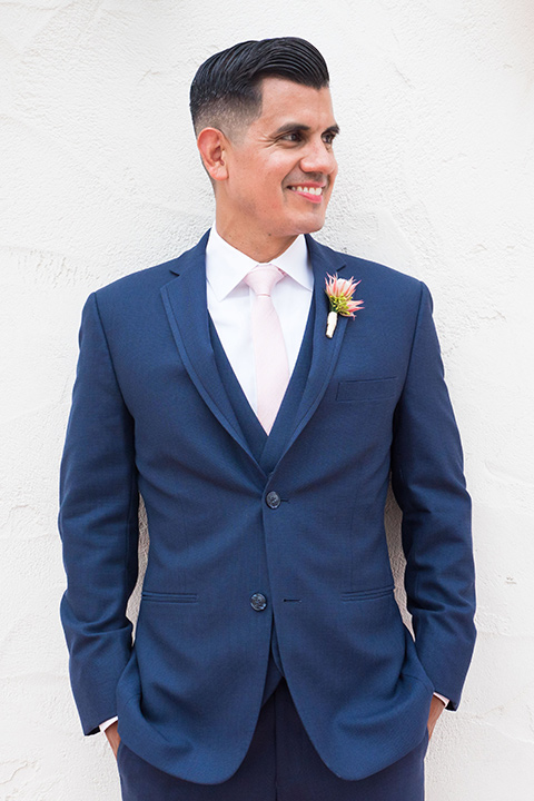 Ole-Hanson-Beach-Club-groom-by-white-wall-in-a-dark-blue-suit-with-a-light-pink-long-tie-and-brown-shoes