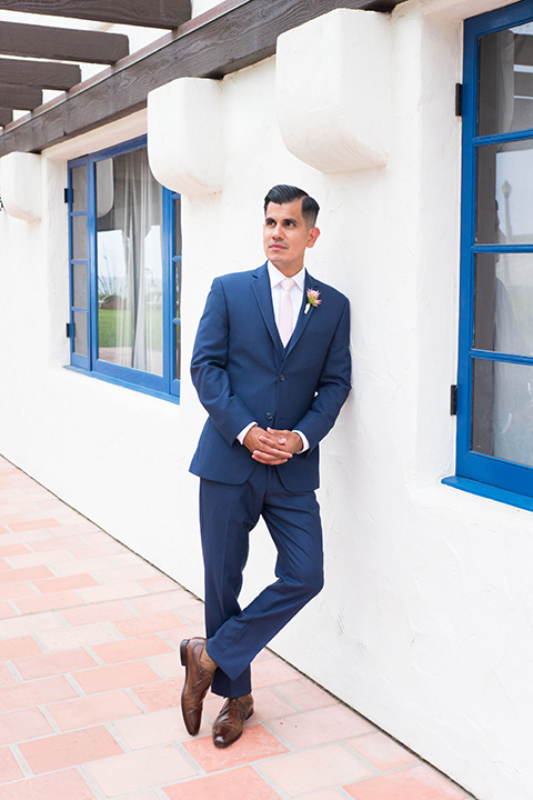 Ole-Hanson-Beach-Club-groom-standing-against-the-wall-in-a-dark-blue-suit-with-a-light-pink-long-tie-and-brown-shoes
