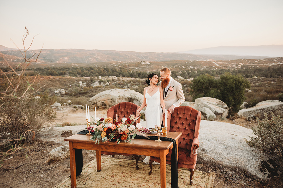 Moonflower-Ranch-Shoot-bride-and-groom-by-table-bride-in-a-form-fitting-lace-gown-with-straps-groom-in-a-tan-jacket-burgundy-pants-and-a-white-floral-long-tie