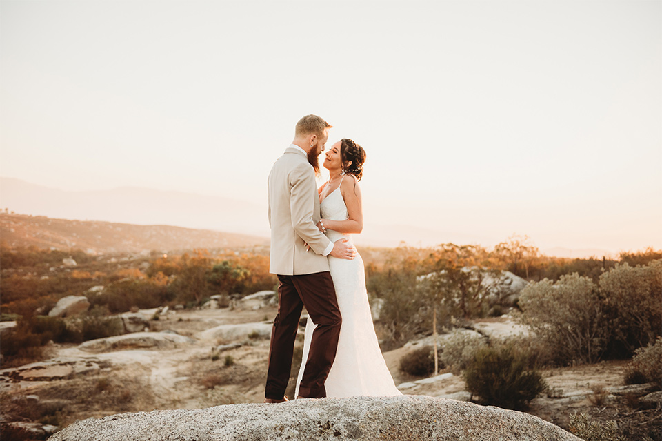 Moonflower-Ranch-Shoot-bride-and-groom-embrace-on-mountain-bride-in-a-form-fitting-lace-gown-with-straps-groom-in-a-tan-jacket-burgundy-pants-and-a-white-floral-long-tie