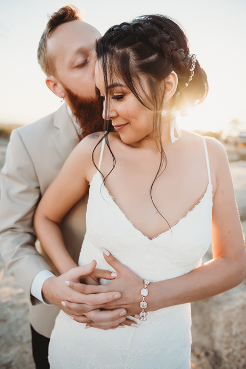 Moonflower-Ranch-Shoot-groom-holding-bride-from-behind-bride-in-a-lace-form-fitting-gown-with-straps-and-v-neck-line-groom-in-a-tan-suit-jacket-with-burgundy-pants-and-floral-necktie