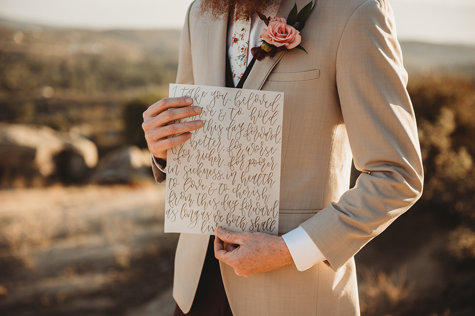 Moonflower-Ranch-Shoot-groom-holding-vows-in-a-tan-jacket-burgundy-pants-and-a-white-floral-long-tie