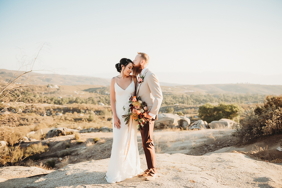 Moonflower-Ranch-Shoot-groom-kissing-bride-on-forehead-bride-in-a-form-fitting-lace-gown-with-straps-groom-in-a-tan-jacket-burgundy-pants-and-a-white-floral-long-tie