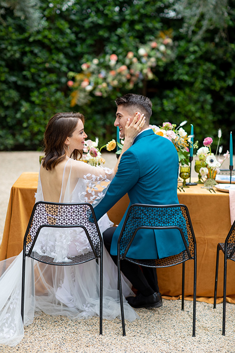 bride and groom at table, bride in a following white gown with a sheer cape and floral design, the groom in a teal tuxedo coat with black pants and a black bow tie