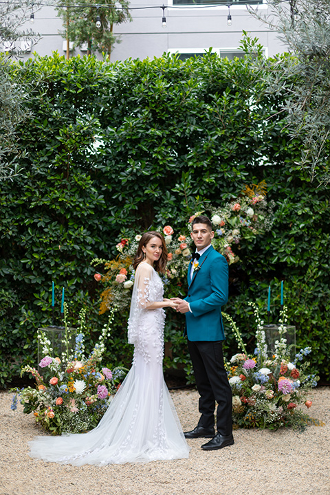 bride and groom at ceremony, bride in a following white gown with a sheer cape and floral design, the groom in a teal tuxedo coat with black pants and a black bow tie 