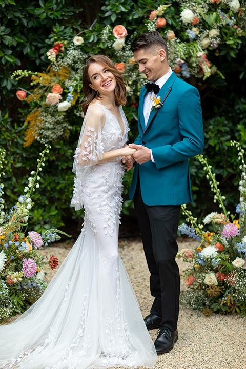 bride and groom at ceremony close up, bride in a following white gown with a sheer cape and floral design, the groom in a teal tuxedo coat with black pants and a black bow tie