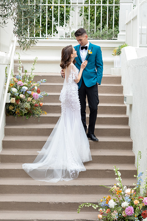 bride and groom standing on steps, bride in a following white gown with a sheer cape and floral design, the groom in a teal tuxedo coat with black pants and a black bow tie 