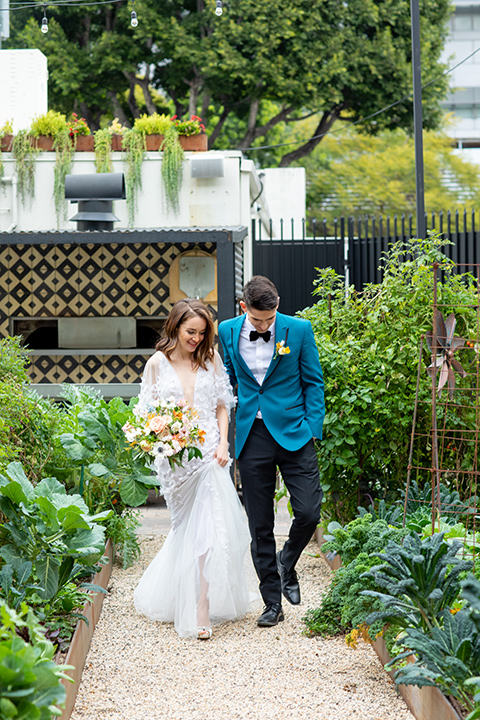 bride and groom walking in the garden, bride in a following white gown with a sheer cape and floral design, the groom in a teal tuxedo coat with black pants and a black bow tie