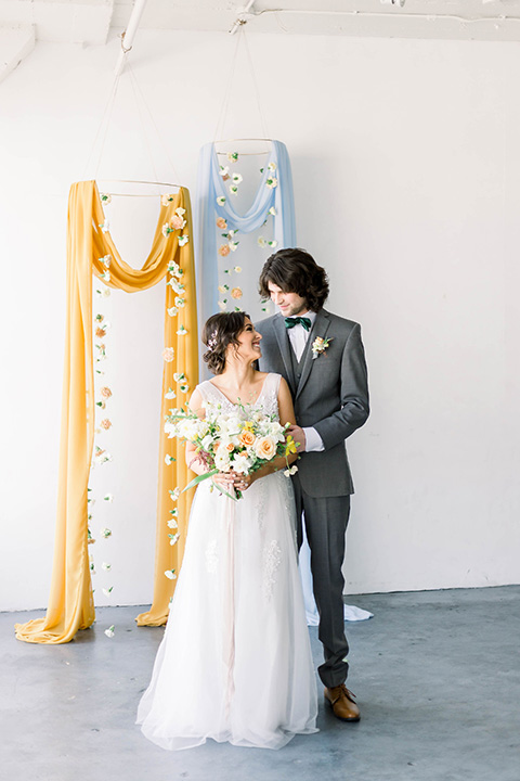 Spring-styled-shoot-bride-and-groom-looking-at-each-other-at-ceremony-space-in-a-bridal-gown-with-a-open-back-and-lace-cap-sleeves-and-soft-tulle-skirt-groom-is-wearing-a-grey-suit-with-a-green-velvet-bow-tie