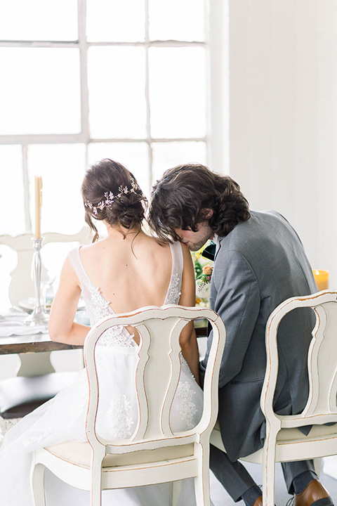 Spring-styled-shoot-bride-and-groom-sitting-at-table-bridal-gown-with-a-open-back-and-lace-cap-sleeves-and-soft-tulle-skirt-the-groom-is-in-a-grey-suit-with-a-green-velvet-bowtie