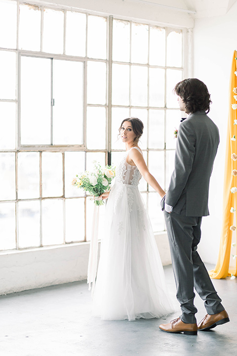 Spring-styled-shoot-bride-and-groom-walking-by-window-the-bride-in-a-gown-with-a-open-back-and-lace-cap-sleeves-and-soft-tulle-skirt-groom-is-wearing-a-grey-suit-with-a-green-velvet-bow-tie