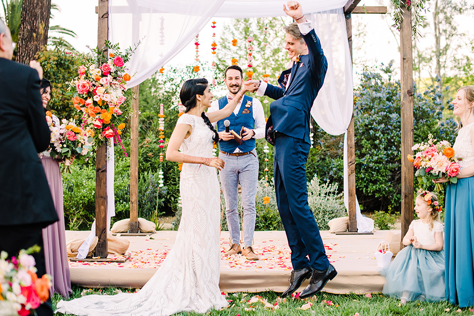 Supriya-and-Chris-wedding-groom-jumping-at-ceremony-the-bride-is-in-a-white-formfitting-gown-with-lace-and-thin-straps-the-groom-in-a-blue-suit-with-pink-bow-tie