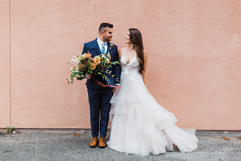 bride in a full tulle ballgown with a crystal and lace bodice and straps, groom in a blue suit with a floral tie and brown shoes