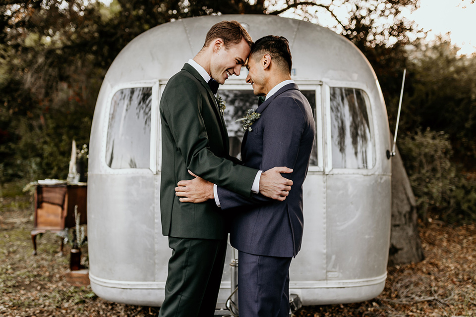 retro-ranch-styled-shoot-couple-in-front-of-airstream-one-groom-wearing-a-dark-blue-suit-with-a-green-velvet-bow-tie-the-other-groom-wearing-a-green-suit-with-a-blue-velvet-bow-tie