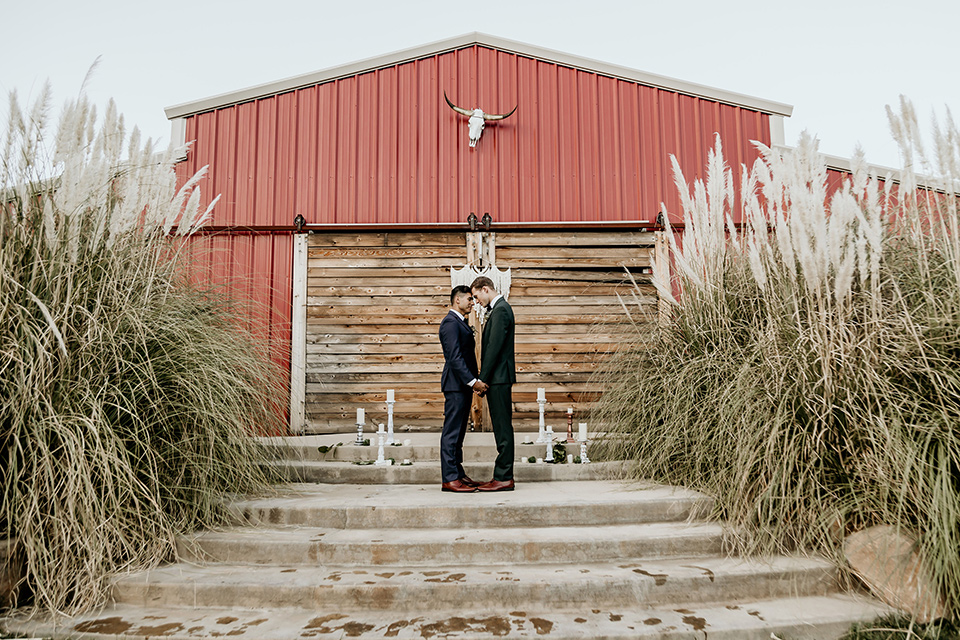 retro-ranch-styled-shoot-grooms-at-ceremony-space-one-groom-wearing-a-dark-blue-suit-with-a-green-velvet-bow-tie-the-other-groom-wearing-a-green-suit-with-a-blue-velvet-bow-tie