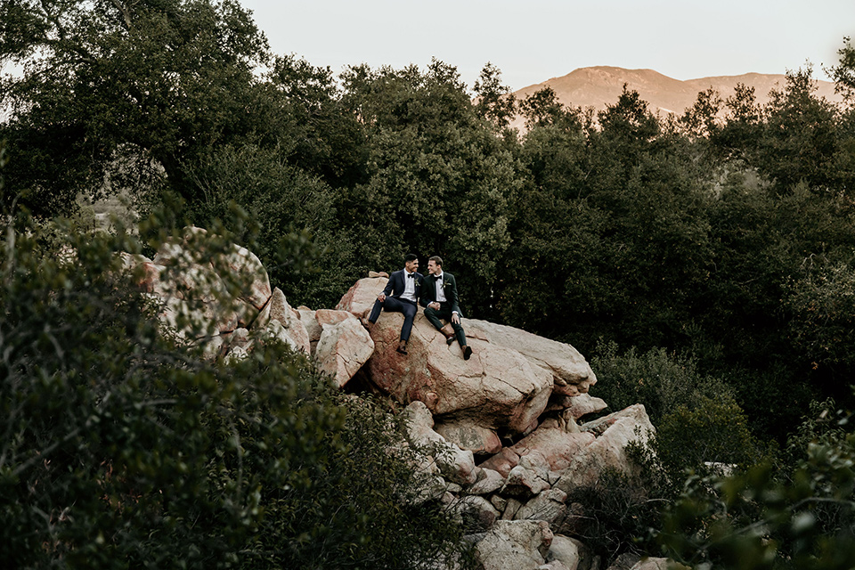 retro-ranch-styled-shoot-grooms-on-rock-one-groom-wearing-a-dark-blue-suit-with-a-green-velvet-bow-tie-the-other-groom-wearing-a-green-suit-with-a-blue-velvet-bow-tie