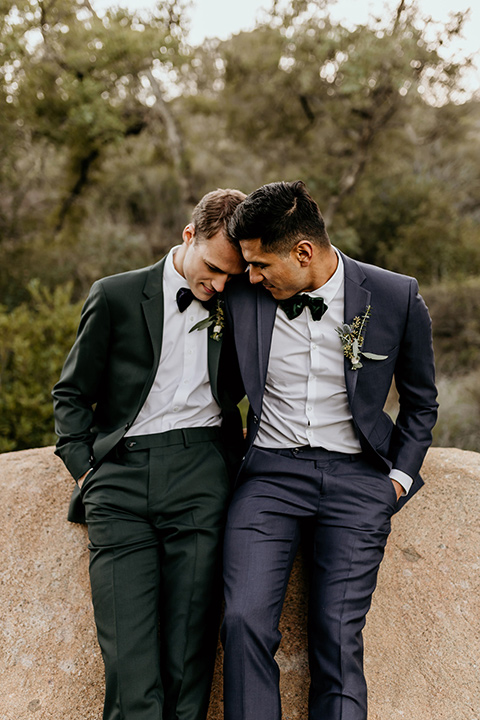 retro-ranch-styled-shoot-grooms-resting-on-rock-together-one-groom-wearing-a-dark-blue-suit-with-a-green-velvet-bow-tie-and-the-other-wearing-a-green-suit-with-a-blue-velvet-bow-tie