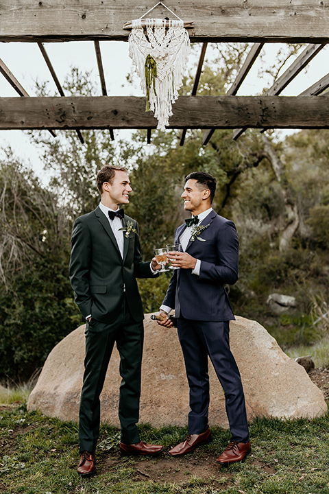 retro-ranch-styled-shoot-grooms-under-macrame-one-groom-wearing-a-dark-blue-suit-with-a-green-velvet-bow-tie-and-the-other-wearing-a-green-suit-with-a-blue-velvet-bow-tie