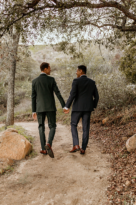 retro-ranch-styled-shoot-grooms-walking-away-one-groom-wearing-a-dark-blue-suit-with-a-green-velvet-bow-tie-and-the-other-wearing-a-green-suit-with-a-blue-velvet-bow-tie