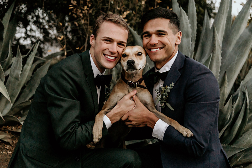 retro-ranch-styled-shoot-grooms-with-dog-one-groom-wearing-a-dark-blue-suit-with-a-green-velvet-bow-tie-the-other-groom-wearing-a-green-suit-with-a-blue-velvet-bow-tie