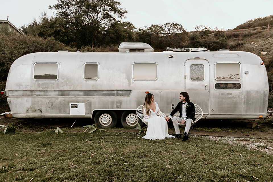 The-Retro-Ranch-Shoot-bride-and-groom-in-front-of-trailer-bride-in-a-boho-gown-with-long-lace-sleeves-and-groom-in-a-velvet-tuxedo-jacket-in-black-with-light-grey-pants