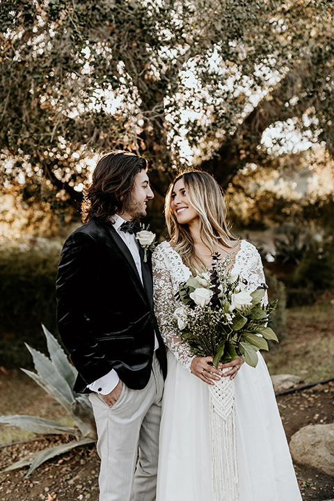 The-Retro-Ranch-Shoot-bride-and-groom-looking-at-each-other-outside-bride-in-a-boho-gown-with-lace-sleeves-and-groom-in-a-black-velvet-coat-with-grey-pants