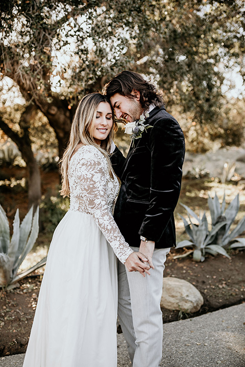 The-Retro-Ranch-Shoot-bride-and-groom-touching-heads-bride-wearing-a-flowing-gown-with-lace-long-sleeves-groom-in-a-black-velvet-coat-with-light-grey-pants