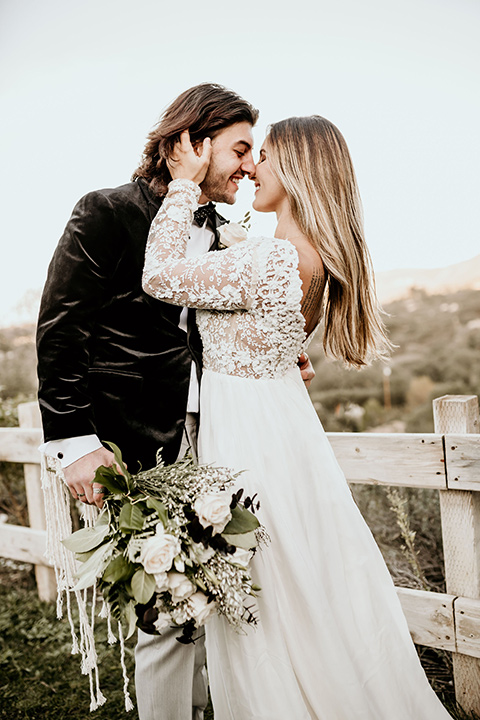 The-Retro-Ranch-Shoot-bride-holding-grooms-face-bride-in-a-boho-gown-with-lace-sleeves-and-groom-in-a-black-velvet-coat-with-grey-pants