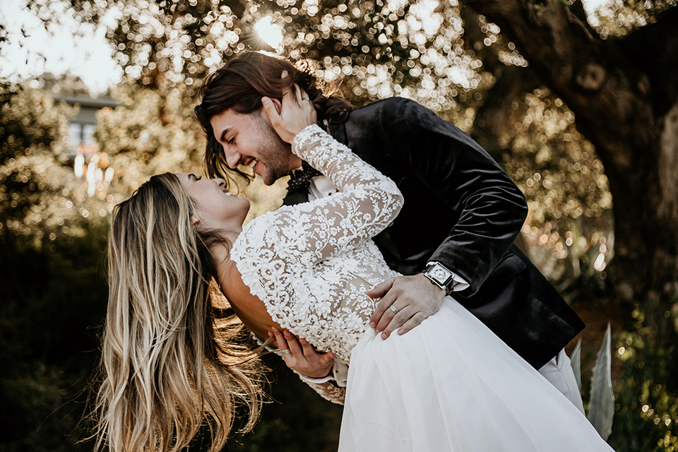 The-Retro-Ranch-Shoot-groom-dipping-bride-boho-gown-with-long-lace-sleeves-and-groom-in-a-velvet-tuxedo-jacket-in-black-with-light-grey-pants