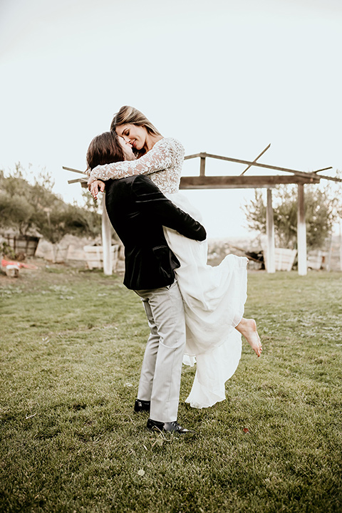 The-Retro-Ranch-Shoot-groom-holding-bride-bride-wearing-a-flowing-gown-with-lace-long-sleeves-groom-in-a-black-velvet-coat-with-light-grey-pants