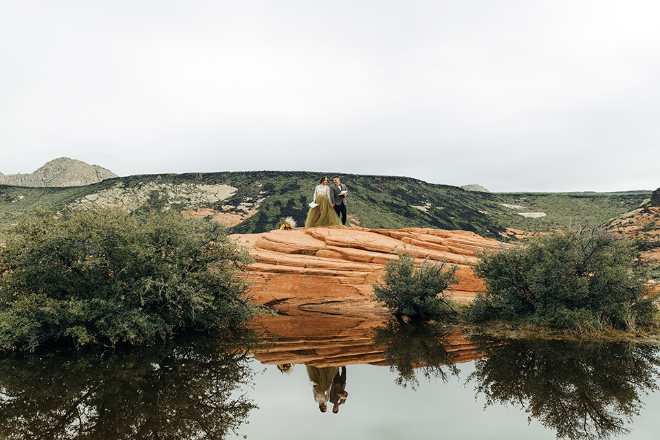 Utah-snow-canyon-shoot-bride-and-groom-by-lake-the-bride-wears-an-olive-green-tulle-skirt-with-an-ivory-lace-long-sleeved-top-and-the-groom-in-a-café-brown-coat-with-a-dark-blue-pant-and-vest-with-a-gold-velvet-bow-tie