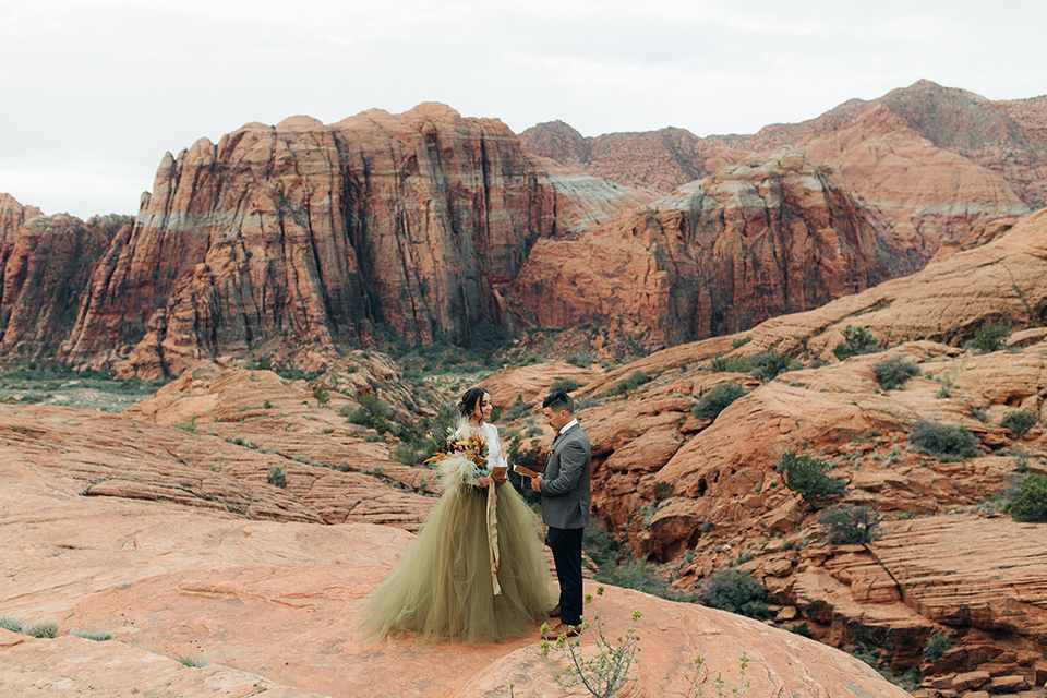 Utah-anow-canyon-shoot-bride-and-groom-facing-each-other-with-vow-book-the-bride-wears-an-olive-green-tulle-skirt-with-an-ivory-lace-long-sleeved-top-and-the-groom-in-a-café-brown-coat-with-a-dark-blue-pant-and-vest-with-a-gold-velvet-bow-tie