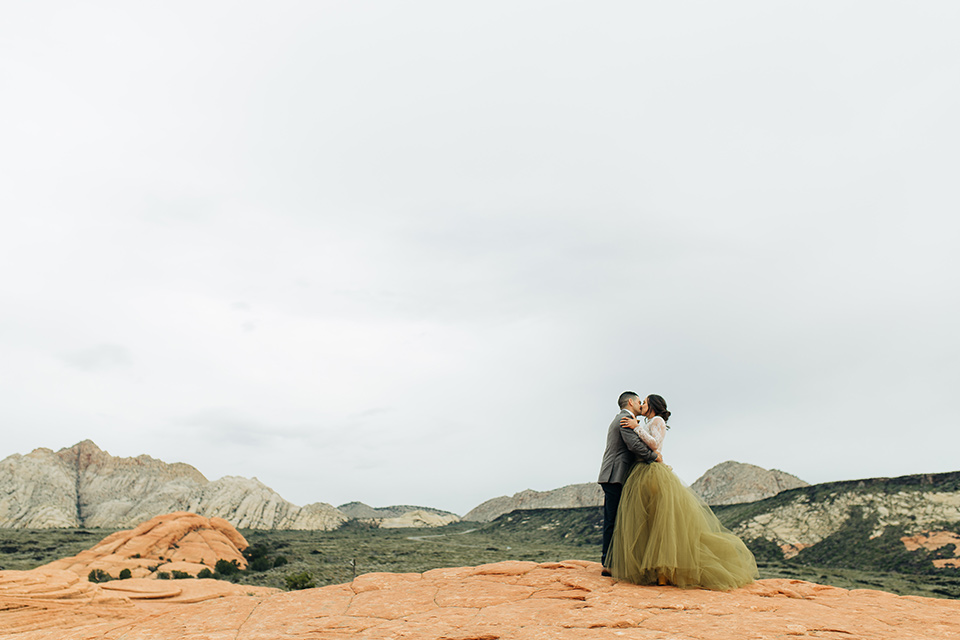 Utah-anow-canyon-shoot-bride-and-groom-holding-eachother-the-bride-wears-an-olive-green-tulle-skirt-with-an-ivory-lace-long-sleeved-top-and-the-groom-in-a-café-brown-coat-with-a-dark-blue-pant-and-vest-with-a-gold-velvet-bow-tie